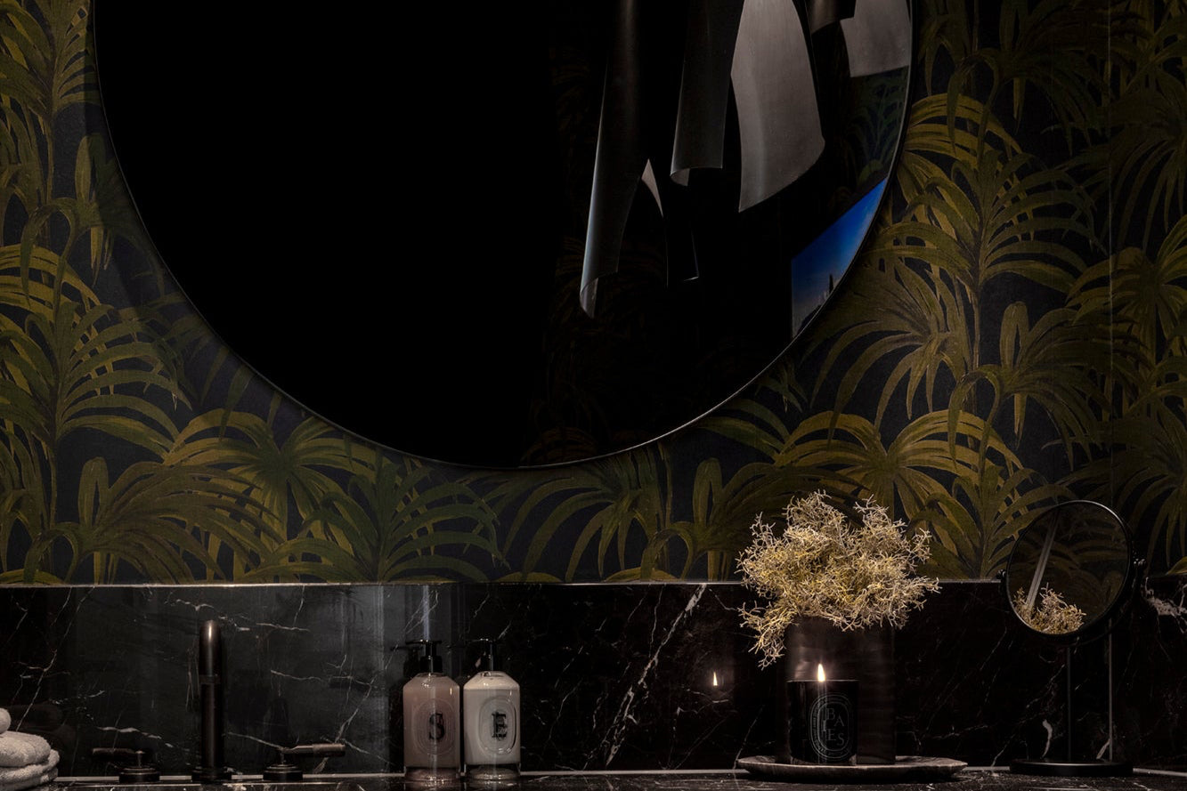 Bathroom with dark green leafy wallpaper, round mirror, marble splashback and decorative elements.