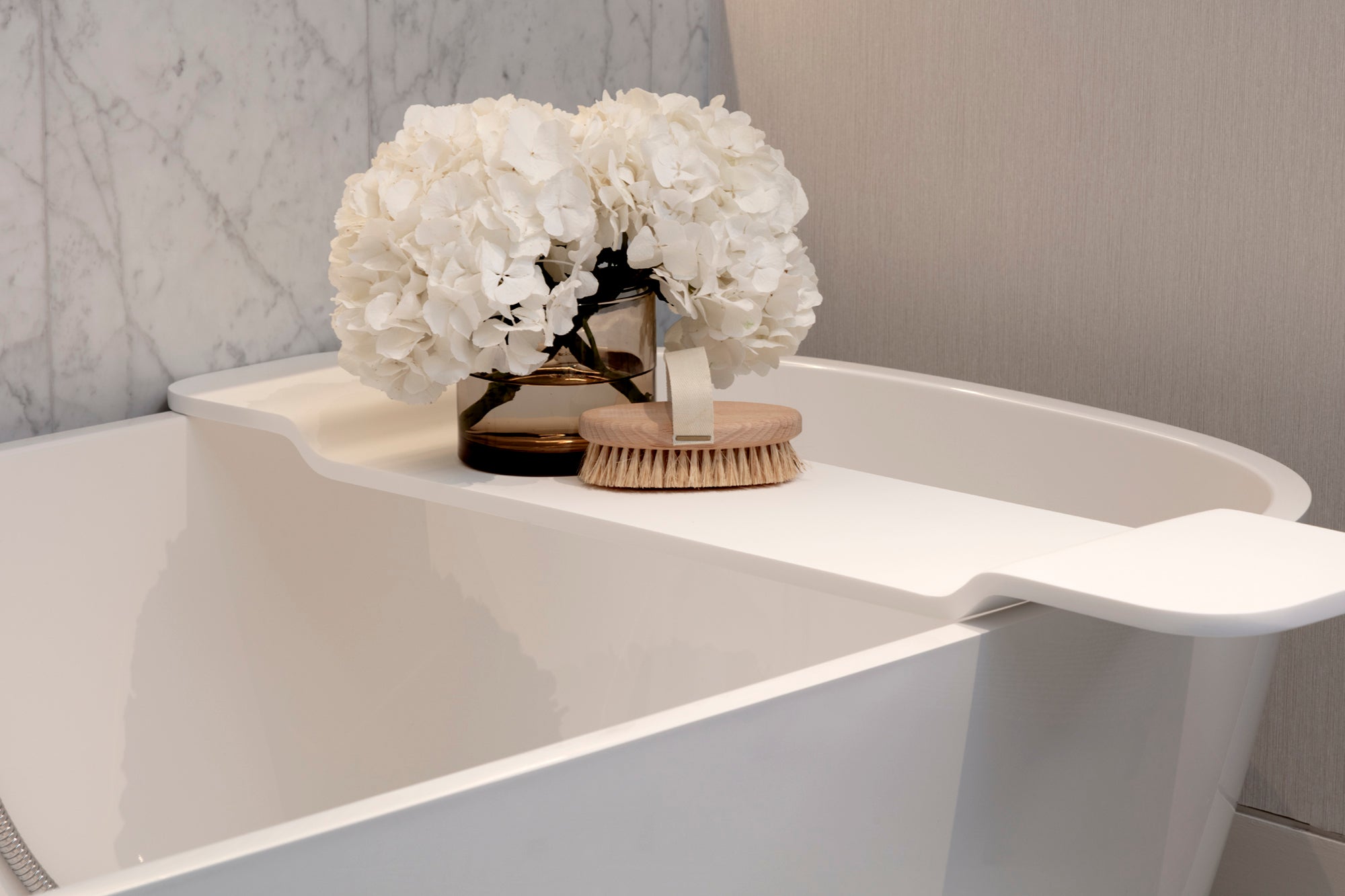White freestanding bathtub with a modern faucet and decorative flowers on a marble wall backdrop. Accessorised by Nineteen Interiors.
