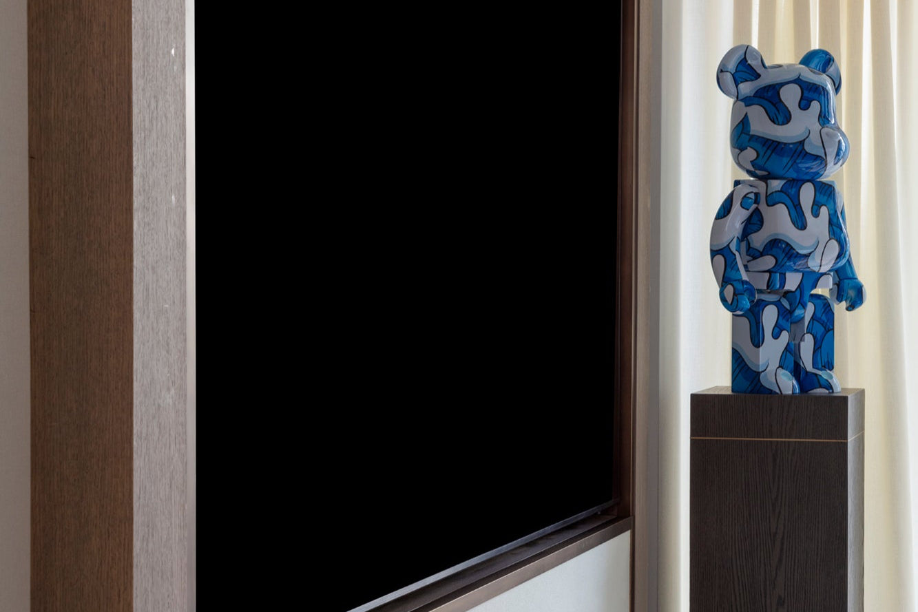Blue and white camouflage bearbrick figure on a timber pedestal next to a large TV screen and bespoke joinery with handmail curtains and voiles.