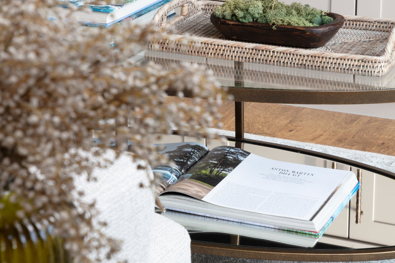 Glass and brass coffee table featuring a luxury classic car book open on top, with a mirrored bottom adding elegance. The neutral tones of the room are accented with subtle punches of colour, creating a refined and stylish space, showcasing high-end luxury interior design