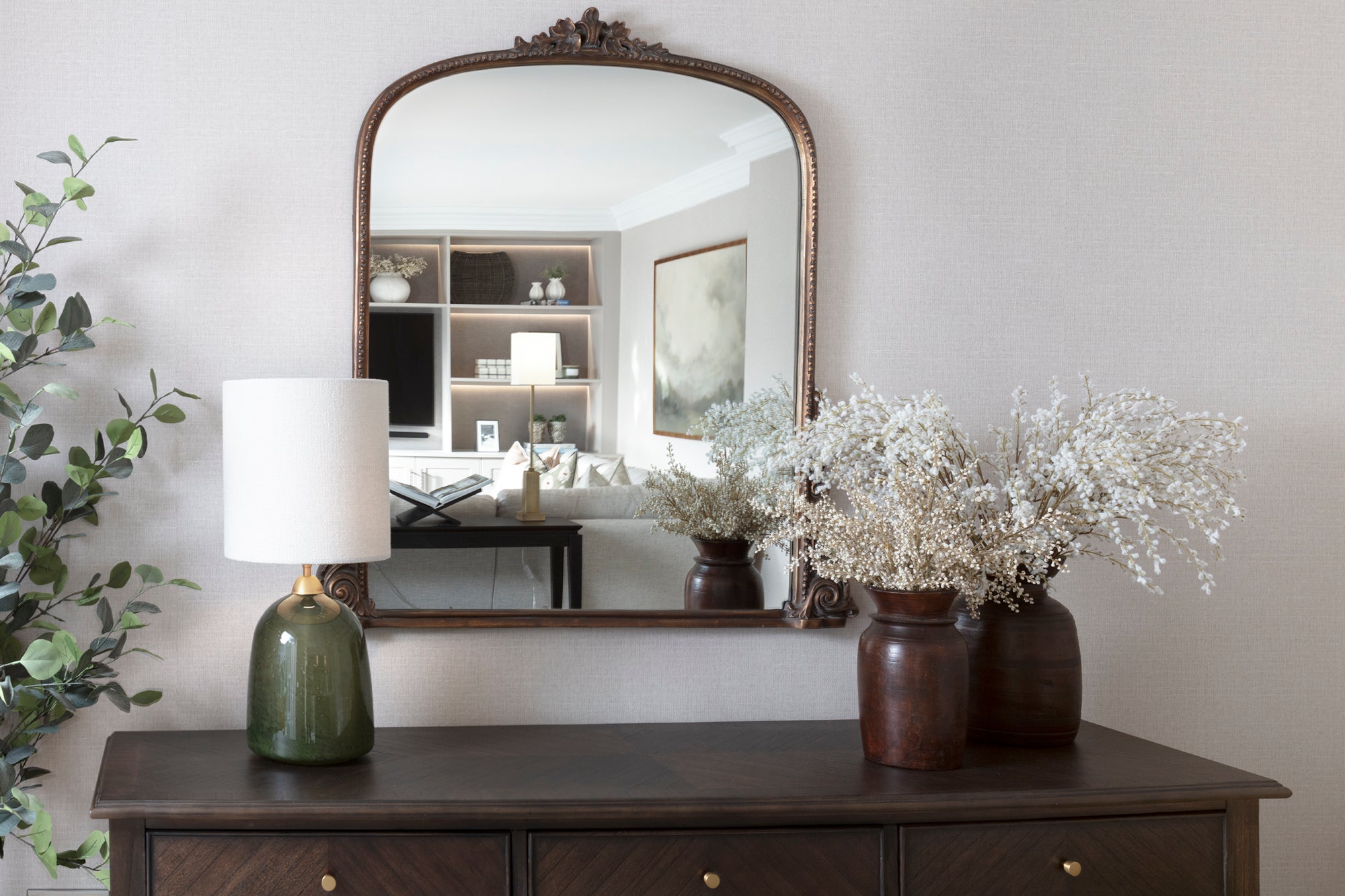 Walnut sideboard with a handmade glass vase and wooden vase featuring faux flowers in a quiet reading space. Custom wallpaper and a luxury mirror enhance the interior design, creating a calm and elegant atmosphere perfect for relaxation